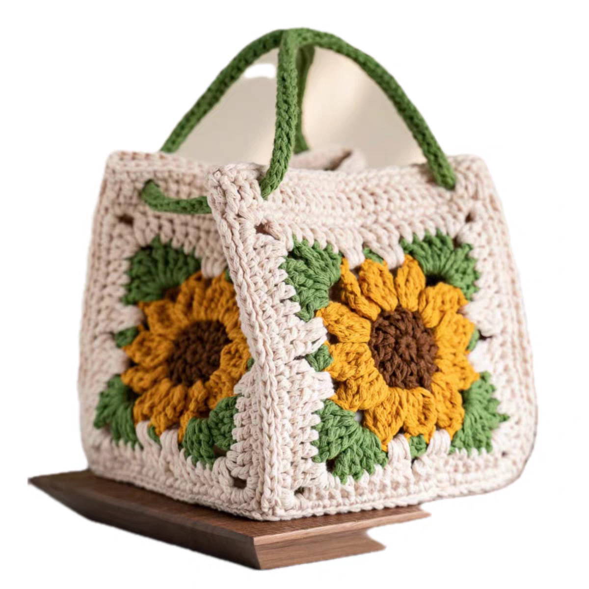 A handmade crocheted tote bag with a cream base, featuring two large yellow sunflower motifs with green leaves and green handles, displayed on a small wooden stand against a white background.