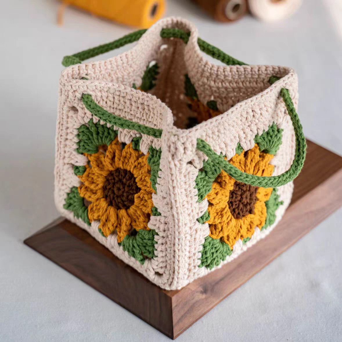 A handmade crocheted storage basket with a sunflower pattern, resting on a dark wooden block against a light gray background.
