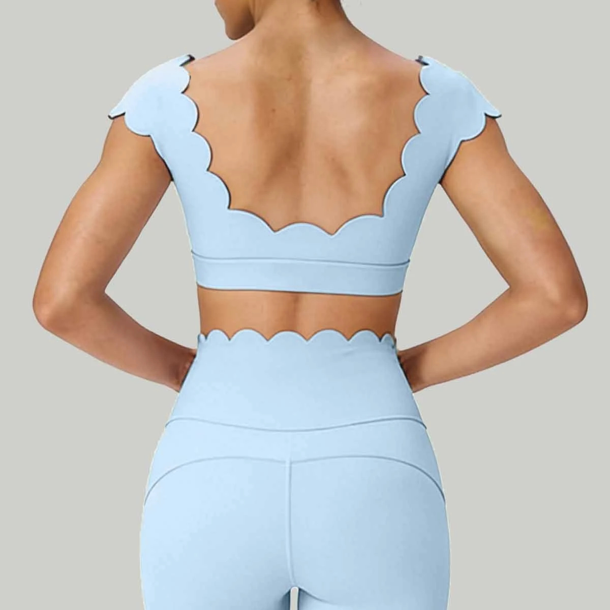 A model shown from the back wearing a light blue matching activewear set with a sports bra featuring scalloped shoulder straps and a high-waisted pair of leggings with a matching scalloped waistband, set against a solid gray background.