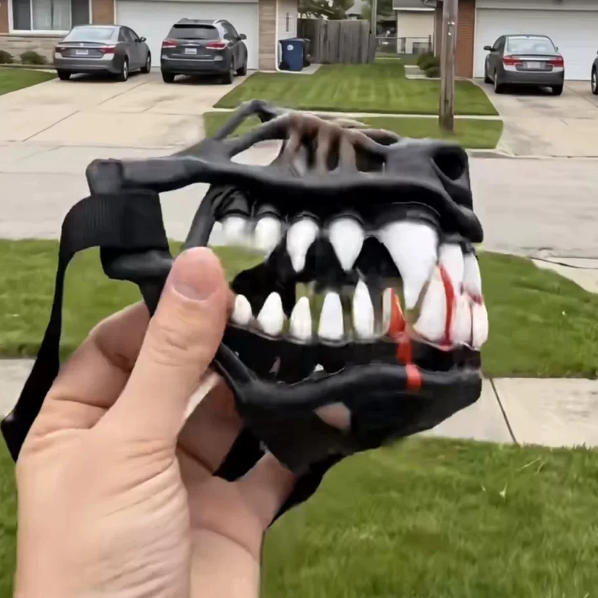 A hand holding a black plastic monster or dinosaur-style mask with white teeth and red blood details, photographed outdoors in front of a suburban lawn and parked cars.