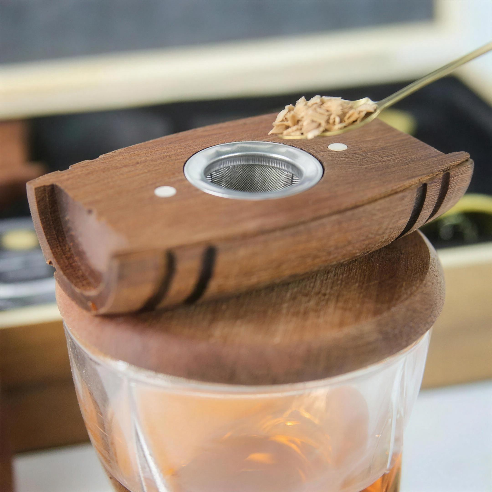 Close-up of a wooden tea infuser lid resting on top of a clear glass teapot with amber tea inside, with a spoon holding loose tea leaves above the infuser opening.