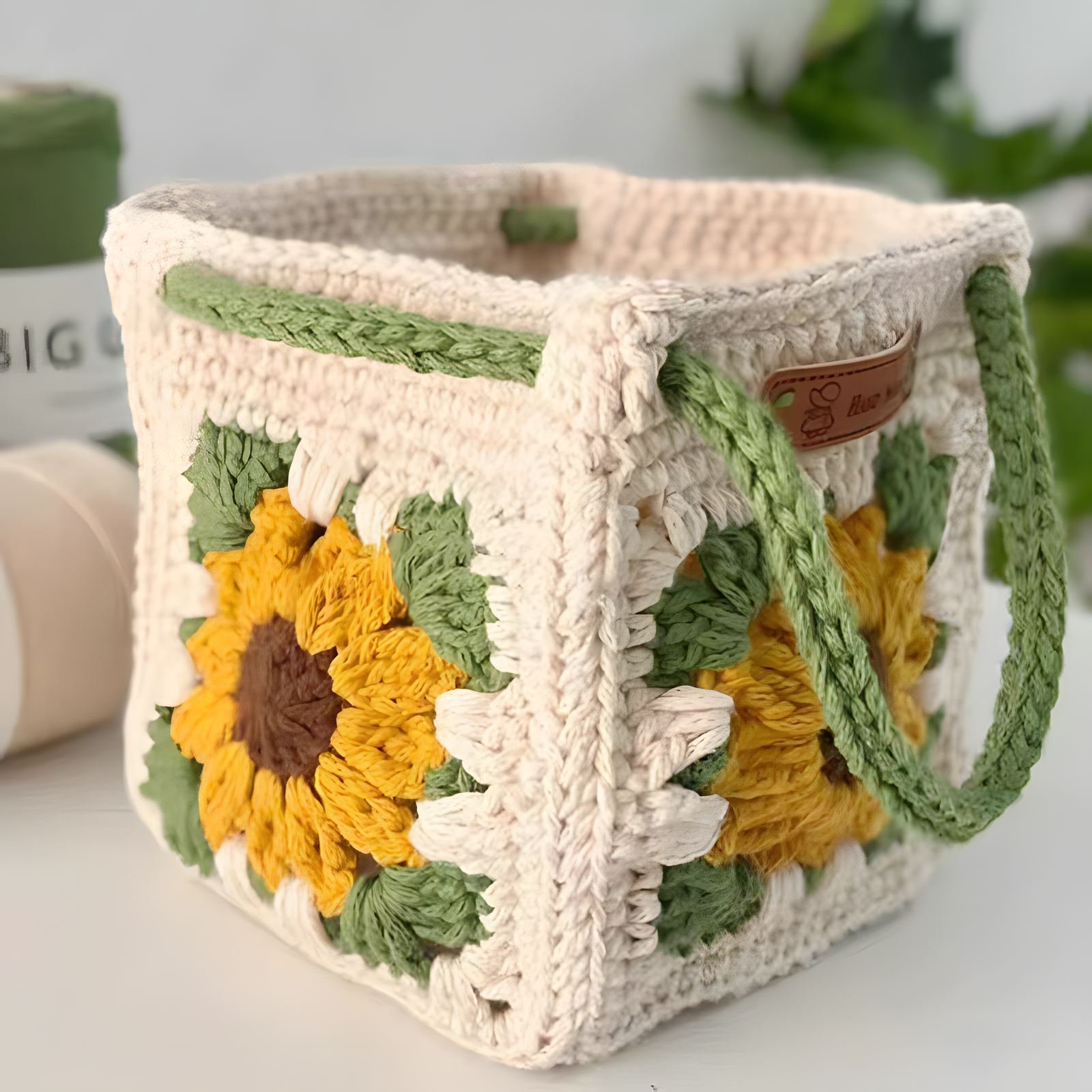 A handmade crocheted basket with a sunflower motif on the front, featuring green handles and a small brown leather brand tag that reads 'S. Bana'.
