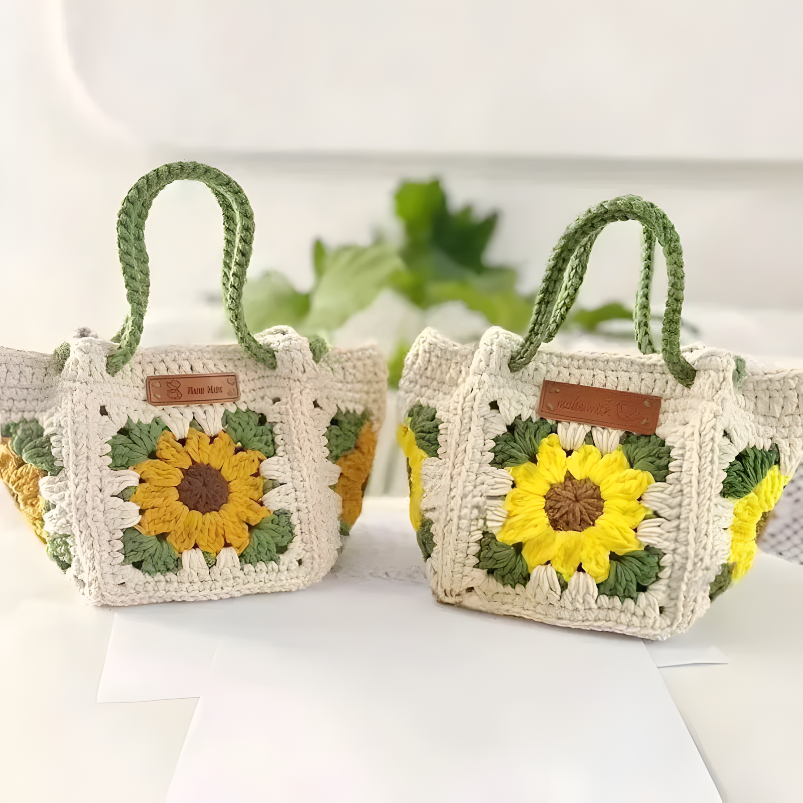 Two handmade crochet tote bags with sunflower designs, featuring off-white crochet bodies, green handles, and yellow and brown sunflower motifs on the front.