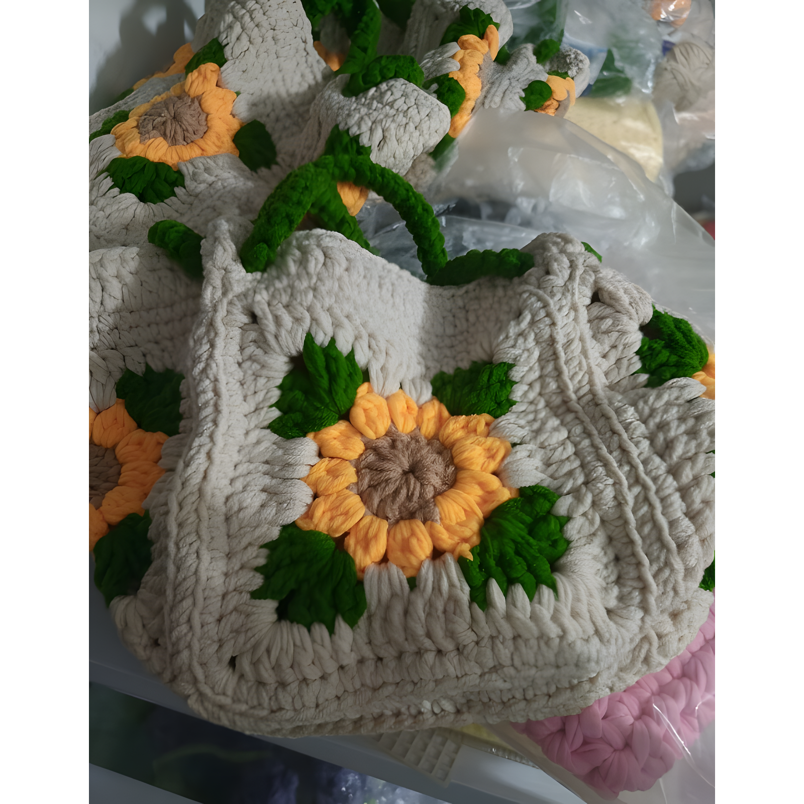 A handmade crocheted tote bag featuring a large yellow and brown sunflower motif with green leaves and dark green handles, displayed alongside other similar bags.